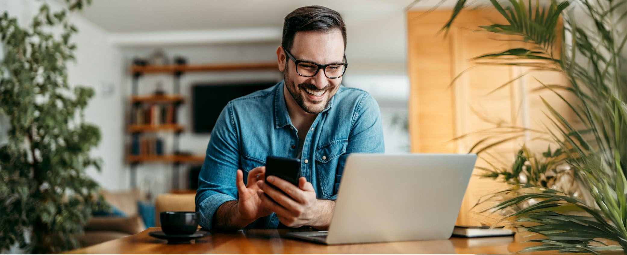 En mand sidder ved et bord og griner med sin telefon og computer