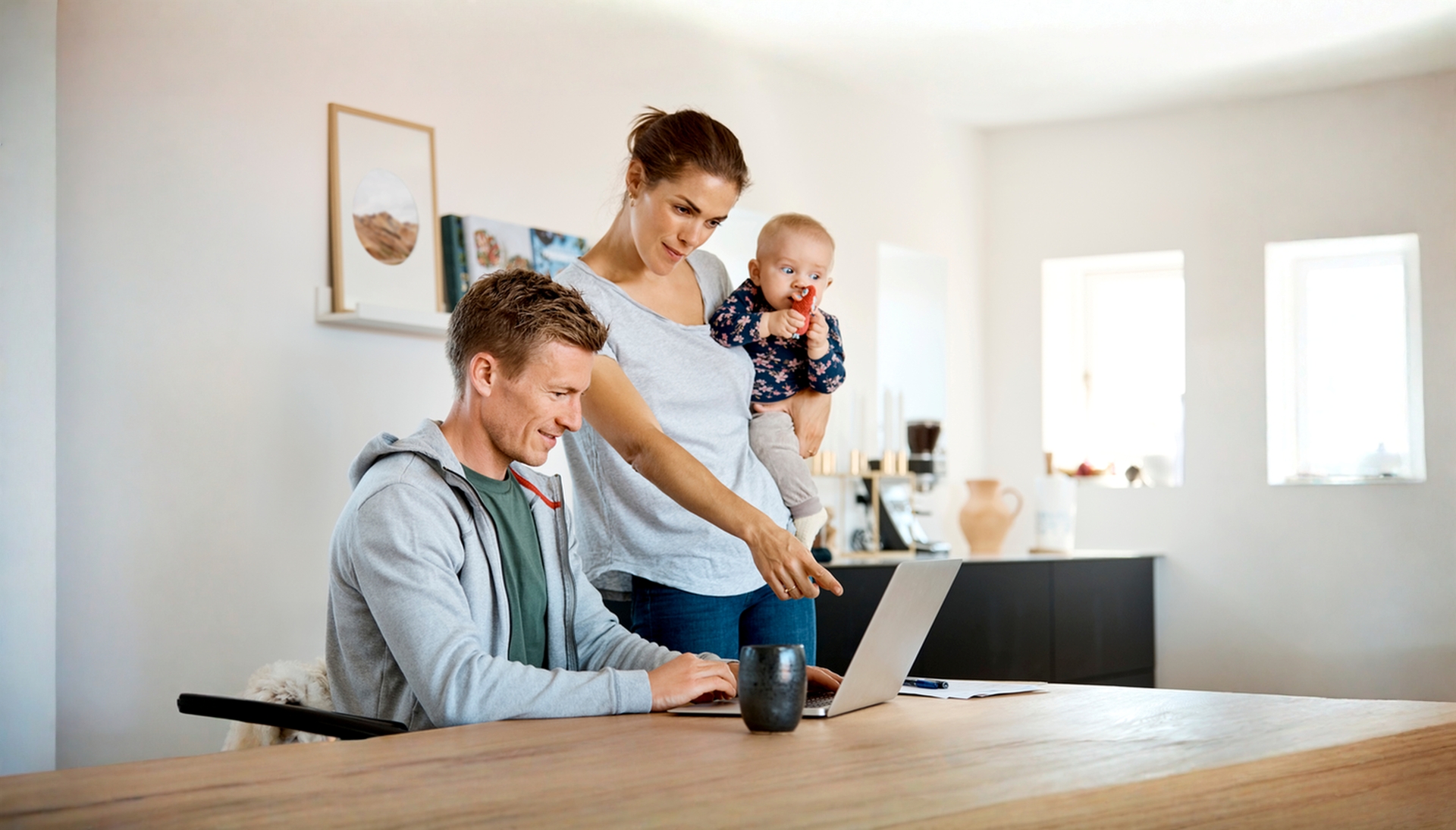 To personer ved en bærbar computer i et hjemmemiljø, mens en baby holdes på armen, hvilket illustrerer digital selvbetjening og håndtering af privatøkonomi.