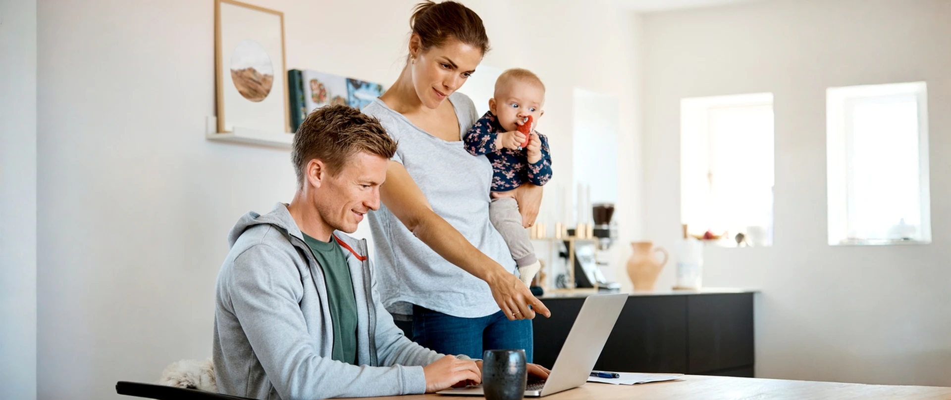 To personer ved en bærbar computer i et hjemmemiljø, mens en baby holdes på armen, hvilket illustrerer digital selvbetjening og håndtering af privatøkonomi.